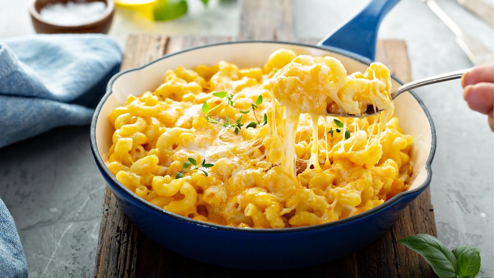 Baked mac and cheese in a blue skillet, topped with melted cheese and herbs, with a spoon lifting a cheesy portion.