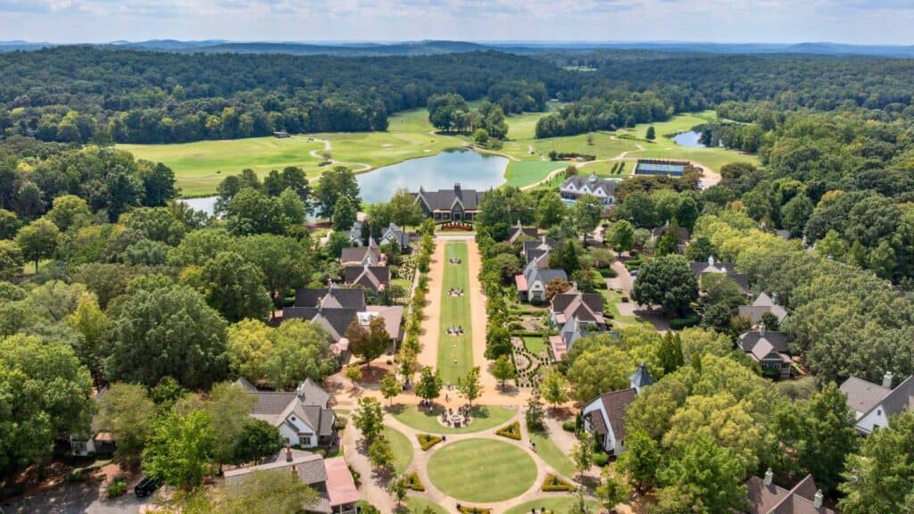 Aerial view of a tree-lined residential neighborhood with a central green area, golf course, lake, and forested surroundings in the background.