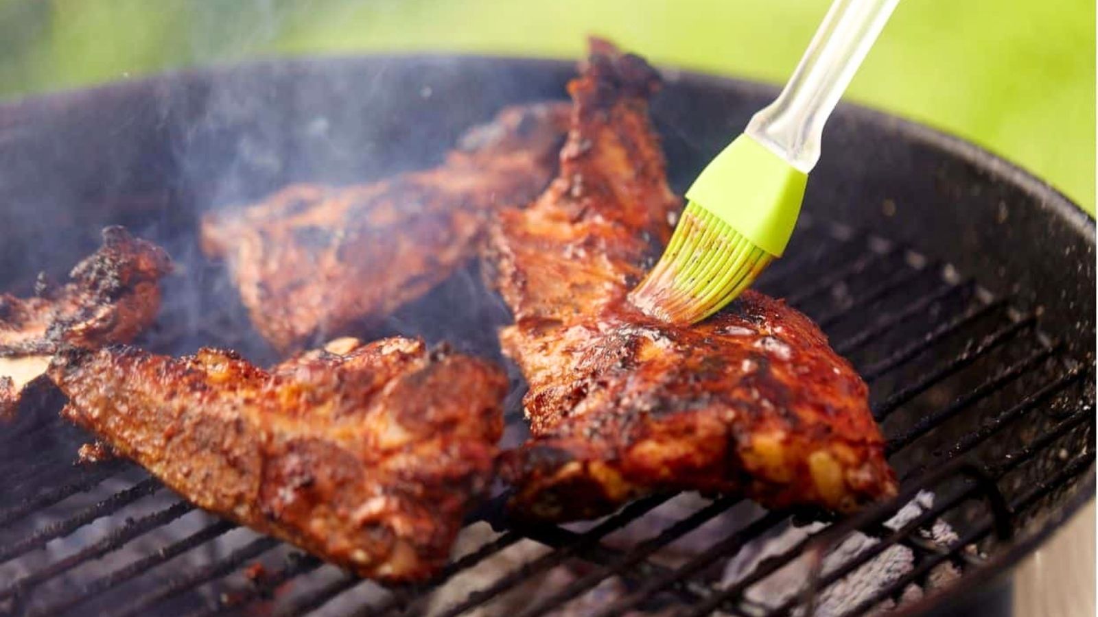 Grilled ribs are being basted with a spicy gochujang sauce using a silicone brush on a barbecue grill, with smoke rising around the meat.