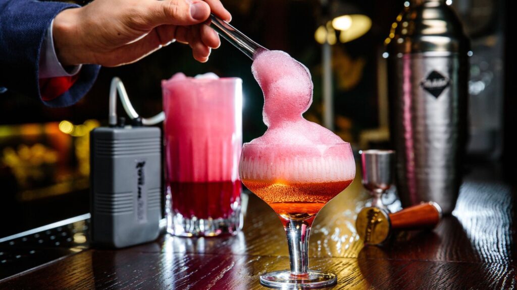 A hand uses tongs to add pink foam to a cocktail in a glass, with bar tools and another pink drink on a wooden bar counter in the background.