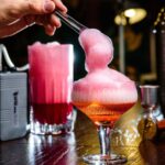 A hand uses tongs to add pink foam to a cocktail in a glass, with bar tools and another pink drink on a wooden bar counter in the background.