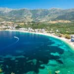 A coastal town with turquoise water, sandy beach, and buildings along the shore—including tempting 5-star hotel bargains—backed by green mountains under a clear blue sky.