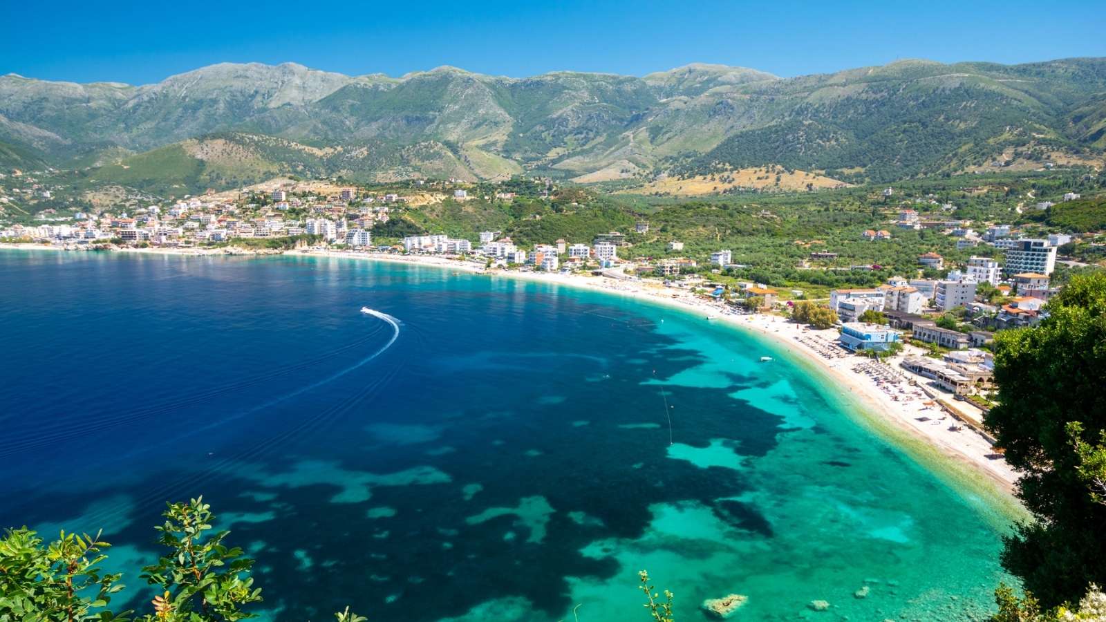 A coastal town with turquoise water, sandy beach, and buildings along the shore—including tempting 5-star hotel bargains—backed by green mountains under a clear blue sky.