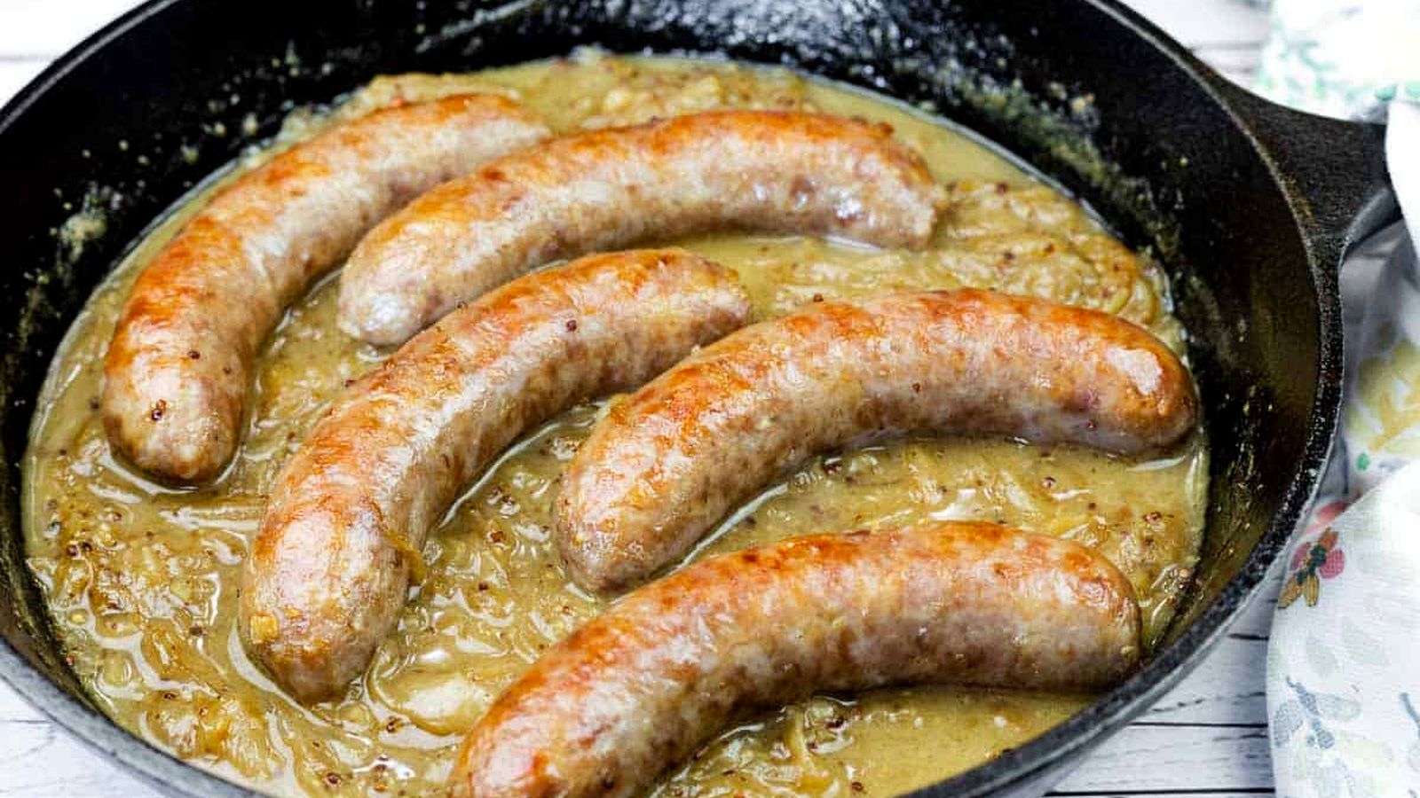 Five browned sausages simmer in onion gravy inside a black cast iron skillet on a light wooden surface.