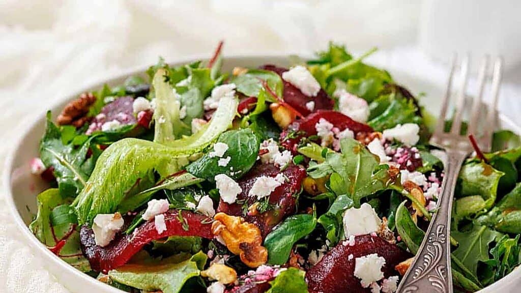 A bowl of mixed greens salad topped with crumbled cheese, sliced beets, and walnuts, with a fork resting on the side.