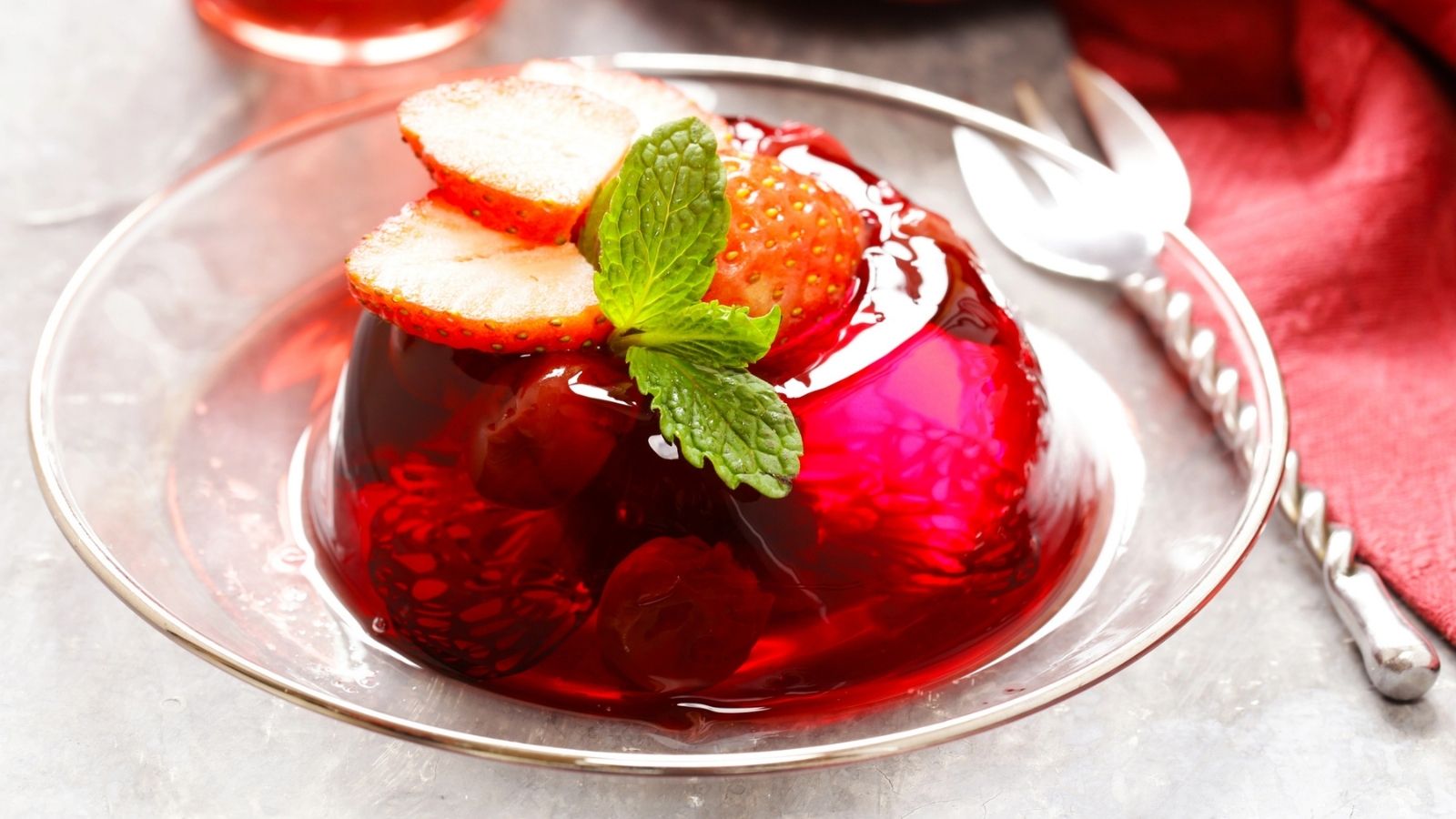 A clear glass plate holds red gelatin dessert topped with strawberry slices and a mint sprig, with a metal dessert fork placed nearby on a light surface.