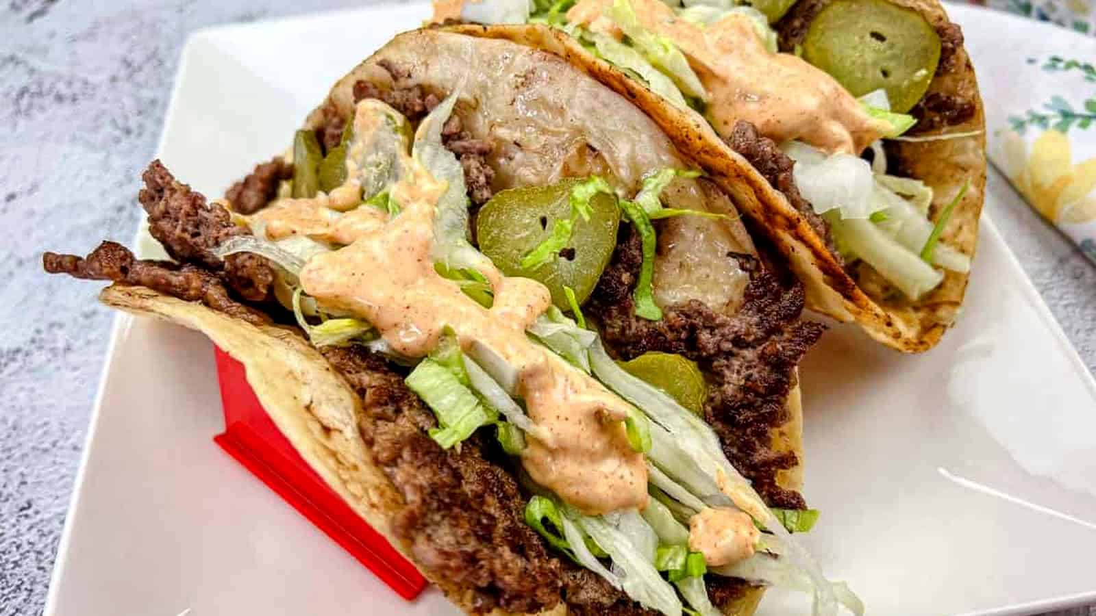 Two Big Mac tacos filled with ground beef, shredded lettuce, and pickles, and drizzled with a creamy sauce, placed on a white plate.