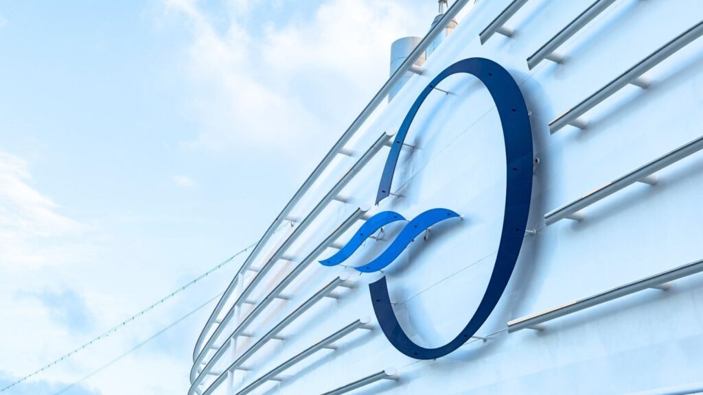 Large blue &ldquo;O&rdquo; logo with two wave shapes is mounted on the side of a white curved structure, likely part of a ship offering Oceania Caribbean sailings, against a blue sky with some clouds.