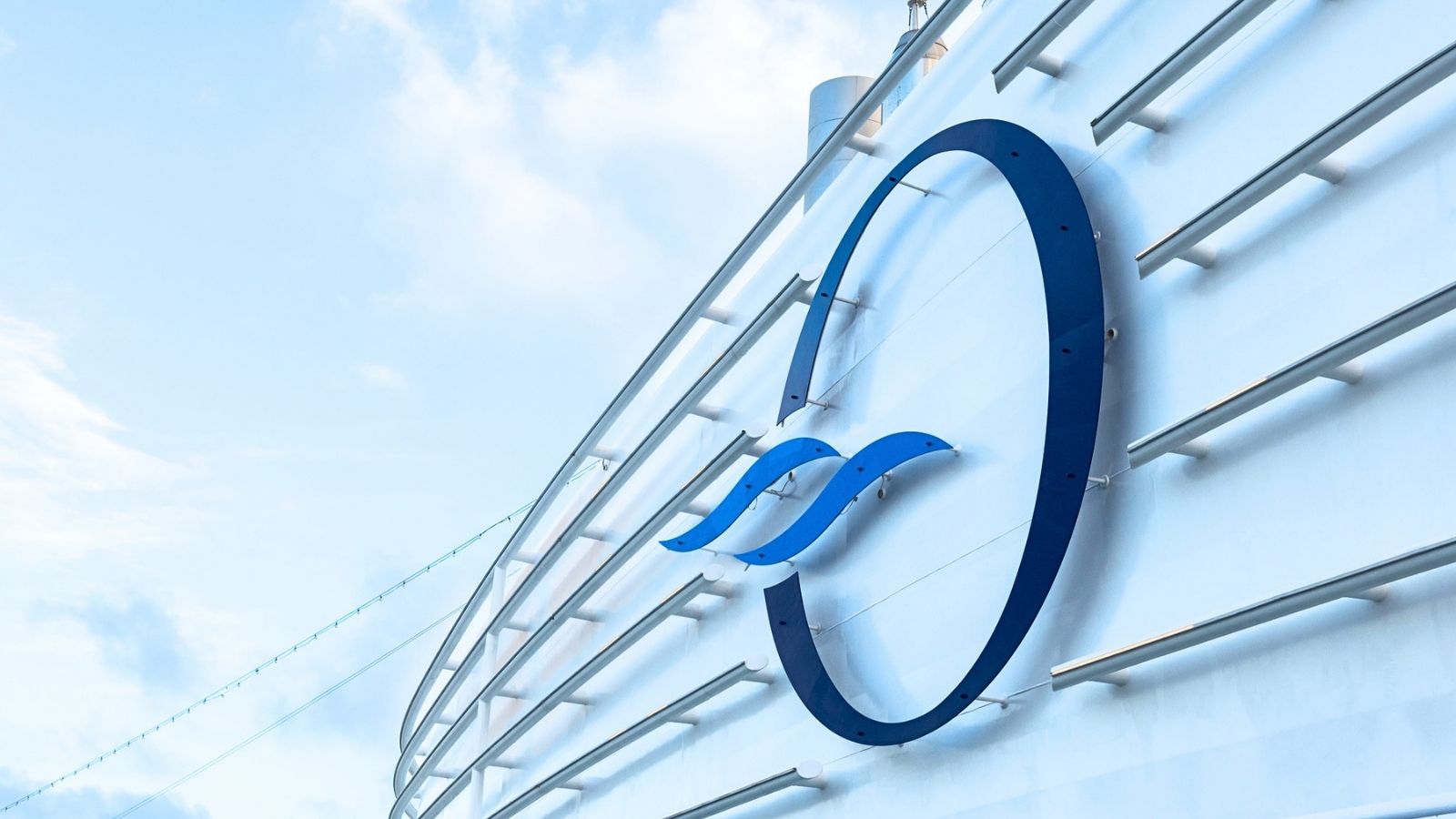 Large blue “O” logo with two wave shapes is mounted on the side of a white curved structure, likely part of a ship offering Oceania Caribbean sailings, against a blue sky with some clouds.