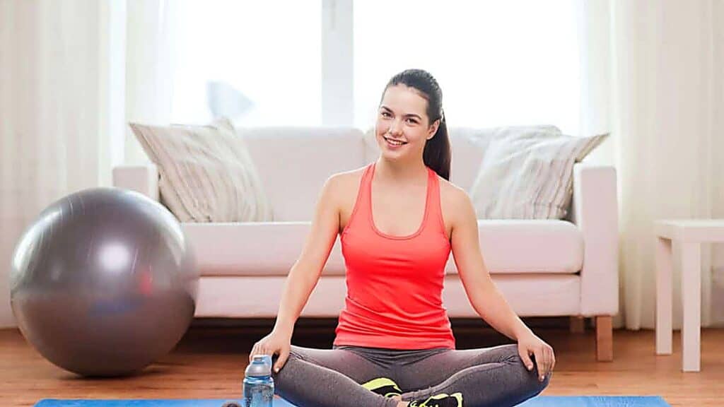 Woman in workout clothes sits cross-legged on a yoga mat indoors, smiling. An exercise ball, water bottle, couch, and white curtains are visible in the background.