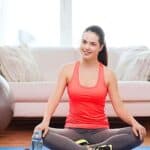 Woman in workout clothes sits cross-legged on a yoga mat indoors, smiling. An exercise ball, water bottle, couch, and white curtains are visible in the background.