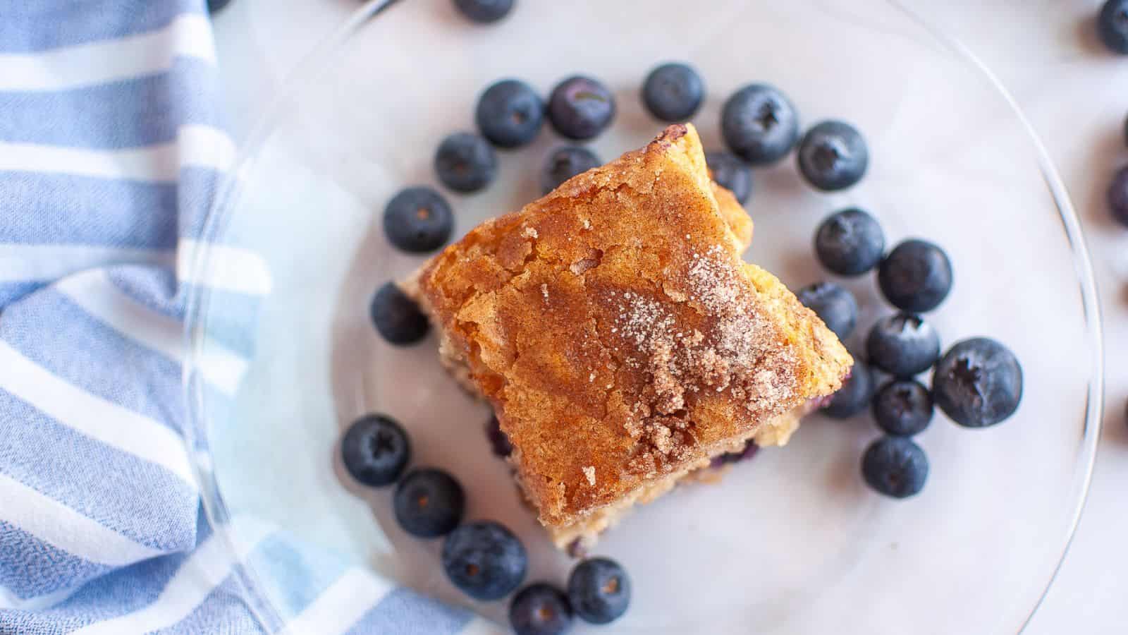 A square piece of blueberry boy bait sits on a clear plate, surrounded by fresh blueberries, next to a blue and white striped cloth.