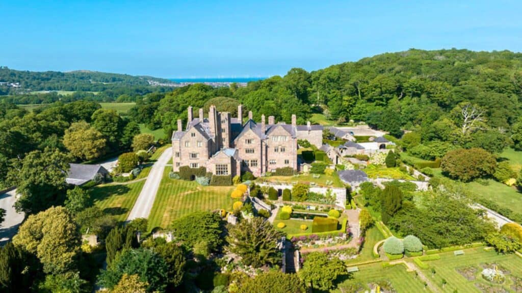 Aerial view of a large, historic stone mansion with multiple chimneys, surrounded by landscaped gardens, trees, and fields under a clear blue sky.