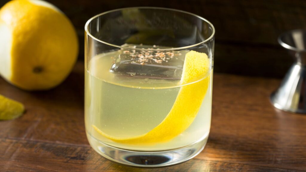 A glass of yellow cocktail with a large ice cube and a lemon peel garnish sits on a wooden surface, with a lemon half and metal jigger in the background.