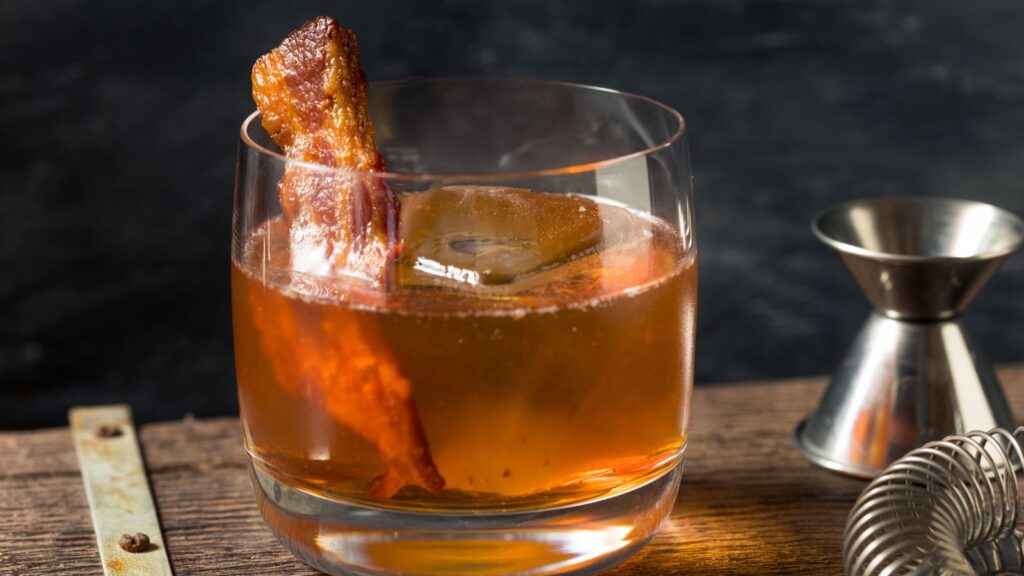 A glass of cocktail with a large ice cube and a strip of crispy bacon garnish, set on a wooden surface with a metal jigger nearby.