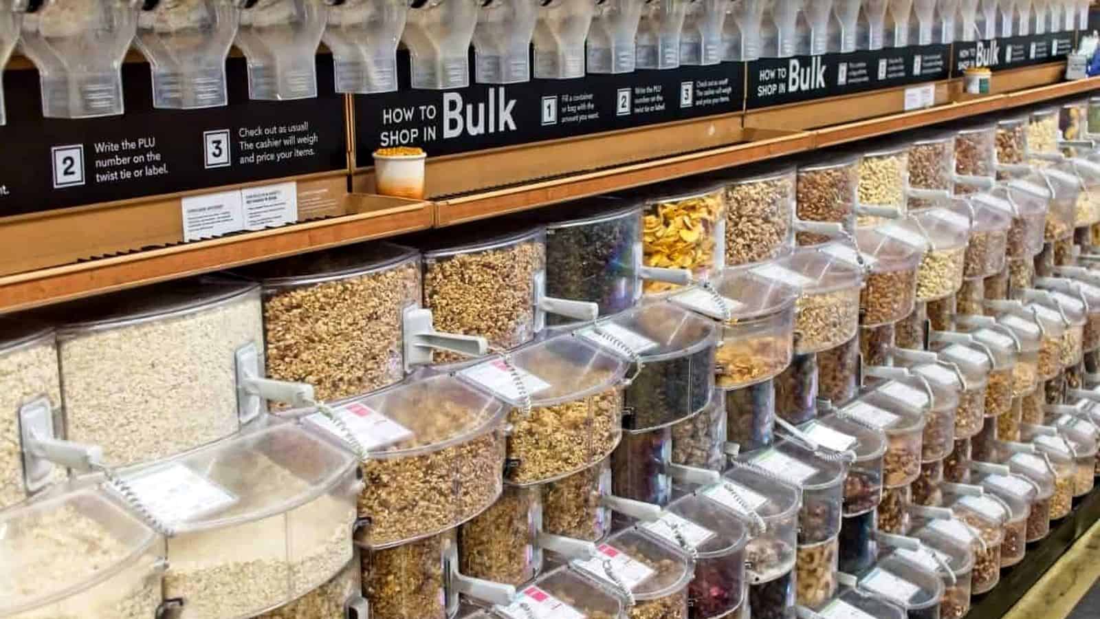 Bulk food dispensers in a store, filled with various grains, nuts, pasta, and dried goods, organized in clear containers with labels and instructions above.