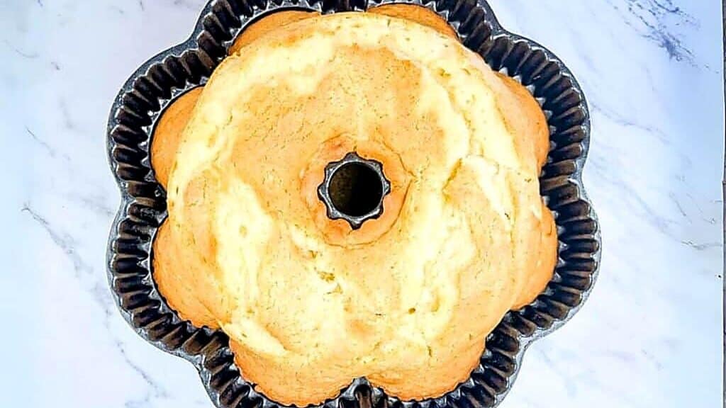 A golden-brown bundt cake rests in a fluted metal baking pan on a white marble surface, viewed from above.