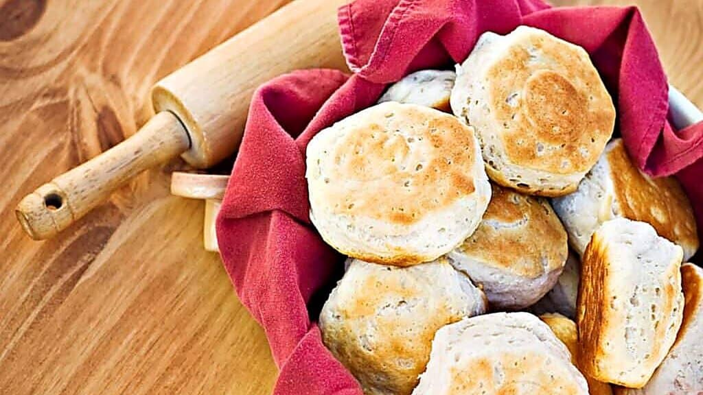 A basket lined with a red cloth holds several golden-brown biscuits, next to a wooden rolling pin on a wooden surface.