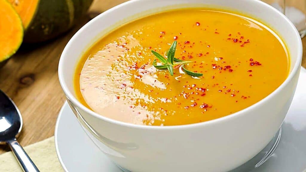 A white bowl of creamy orange soup, garnished with red spices and a sprig of rosemary, sits on a saucer with a spoon nearby.
