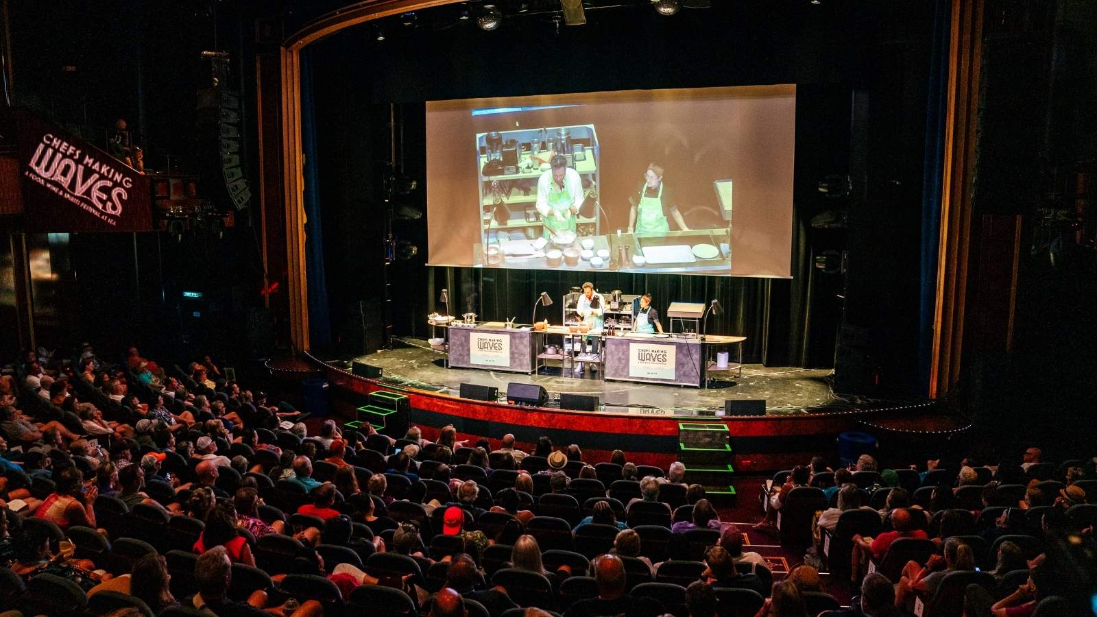 A large audience watches a live cooking demonstration on stage, with two presenters shown on a big screen above them, reminiscent of the lively culinary experiences often featured on Sixthman Cruises.
