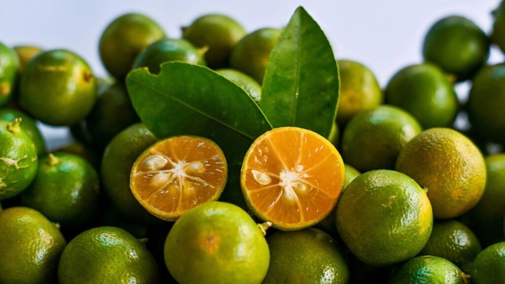 A pile of calamansi limes with two green leaves and one lime cut in half to show its orange interior.