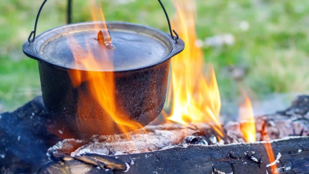 A black metal pot hangs over an open campfire, with orange flames surrounding the pot and logs below, outdoors on grass.