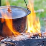 A black metal pot hangs over an open campfire, with orange flames surrounding the pot and logs below, outdoors on grass.