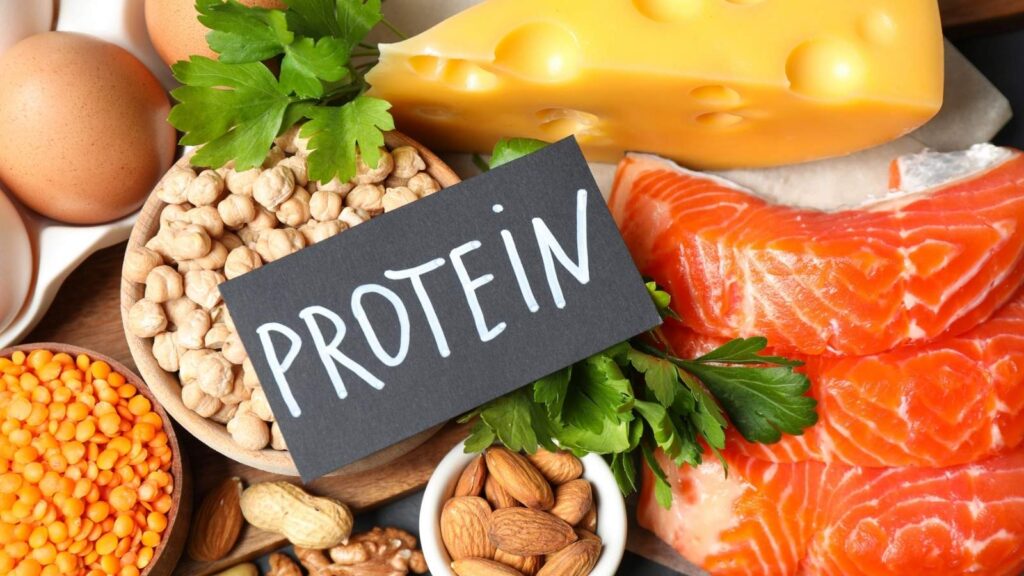 Assorted protein-rich foods including salmon, cheese, eggs, chickpeas, lentils, almonds, and parsley arranged around a card labeled "PROTEIN.