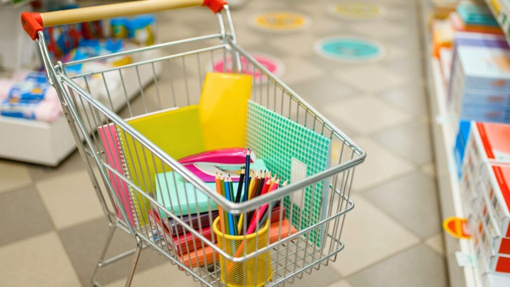 A shopping cart filled with school supplies, including notebooks, folders, colored pencils, and paper, in the aisle of a store with shelves and floor markers visible.