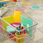 A shopping cart filled with school supplies, including notebooks, folders, colored pencils, and paper, in the aisle of a store with shelves and floor markers visible.