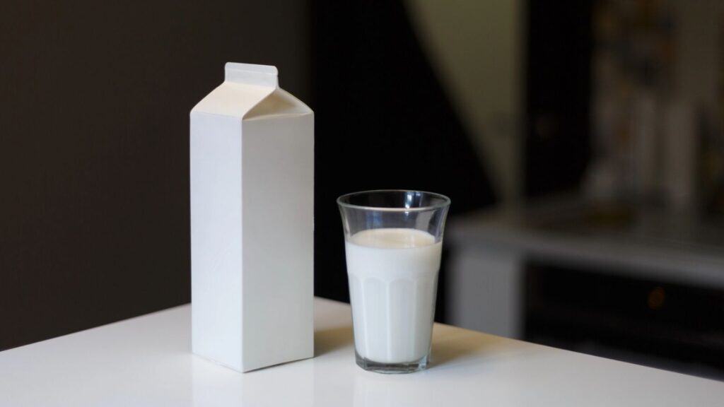 A plain white milk carton and a glass filled with milk are placed on a white surface indoors.