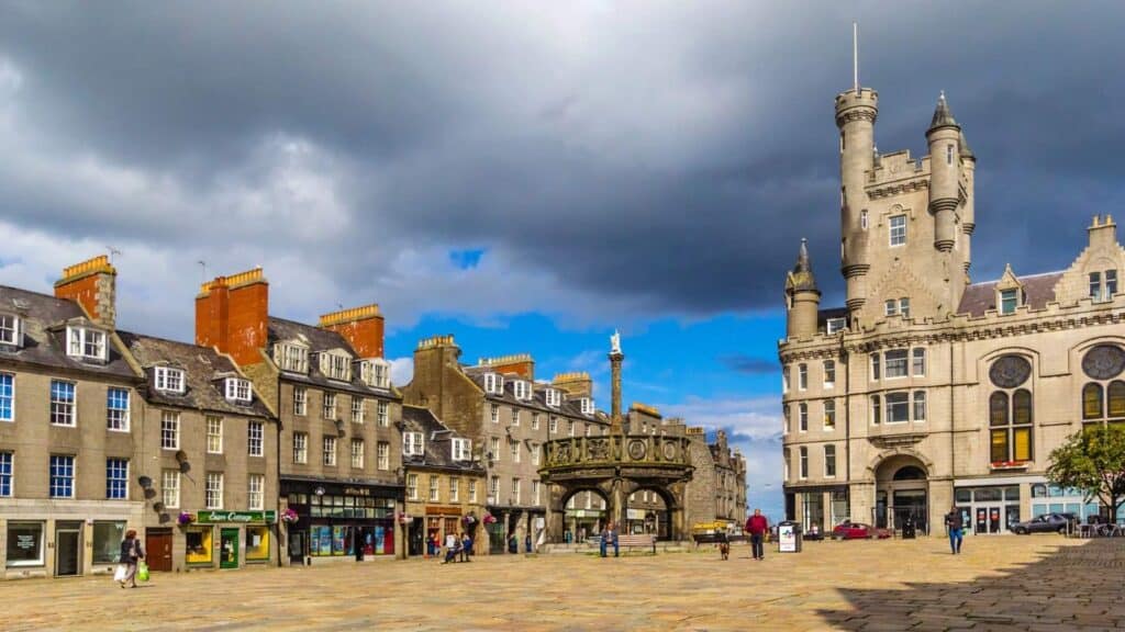 A historic city square with stone buildings, shops, and a prominent clock tower under a cloudy sky, this vibrant plaza is one of the unique travel destinations where people stroll across the open space, soaking in centuries of charm and character.