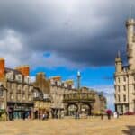 A historic city square with stone buildings, shops, and a prominent clock tower under a cloudy sky, this vibrant plaza is one of the unique travel destinations where people stroll across the open space, soaking in centuries of charm and character.
