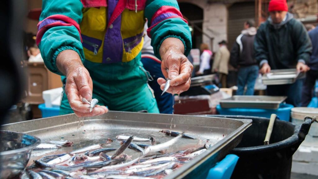 A person in a colorful jacket sorts small fish in a metal tray at a busy outdoor market, capturing the lively scenes travelers might encounter after US flights to Italy, with other people and containers visible in the background.