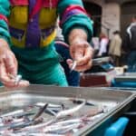 A person in a colorful jacket sorts small fish in a metal tray at a busy outdoor market, capturing the lively scenes travelers might encounter after US flights to Italy, with other people and containers visible in the background.