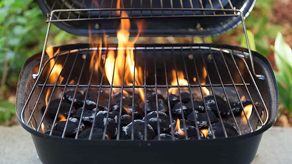 A charcoal grill with flames and burning briquettes beneath the metal grate, outdoors with a blurred green background&mdash;perfect for practicing grilling tips for beginners.