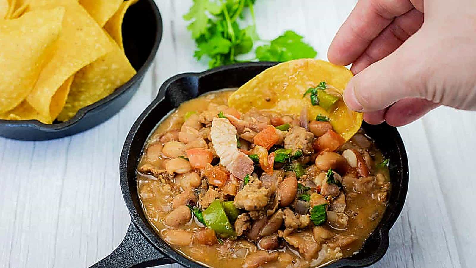 A hand dips a tortilla chip into a small cast-iron skillet filled with Charro Beans With Chorizo, more chips and cilantro are in the background.