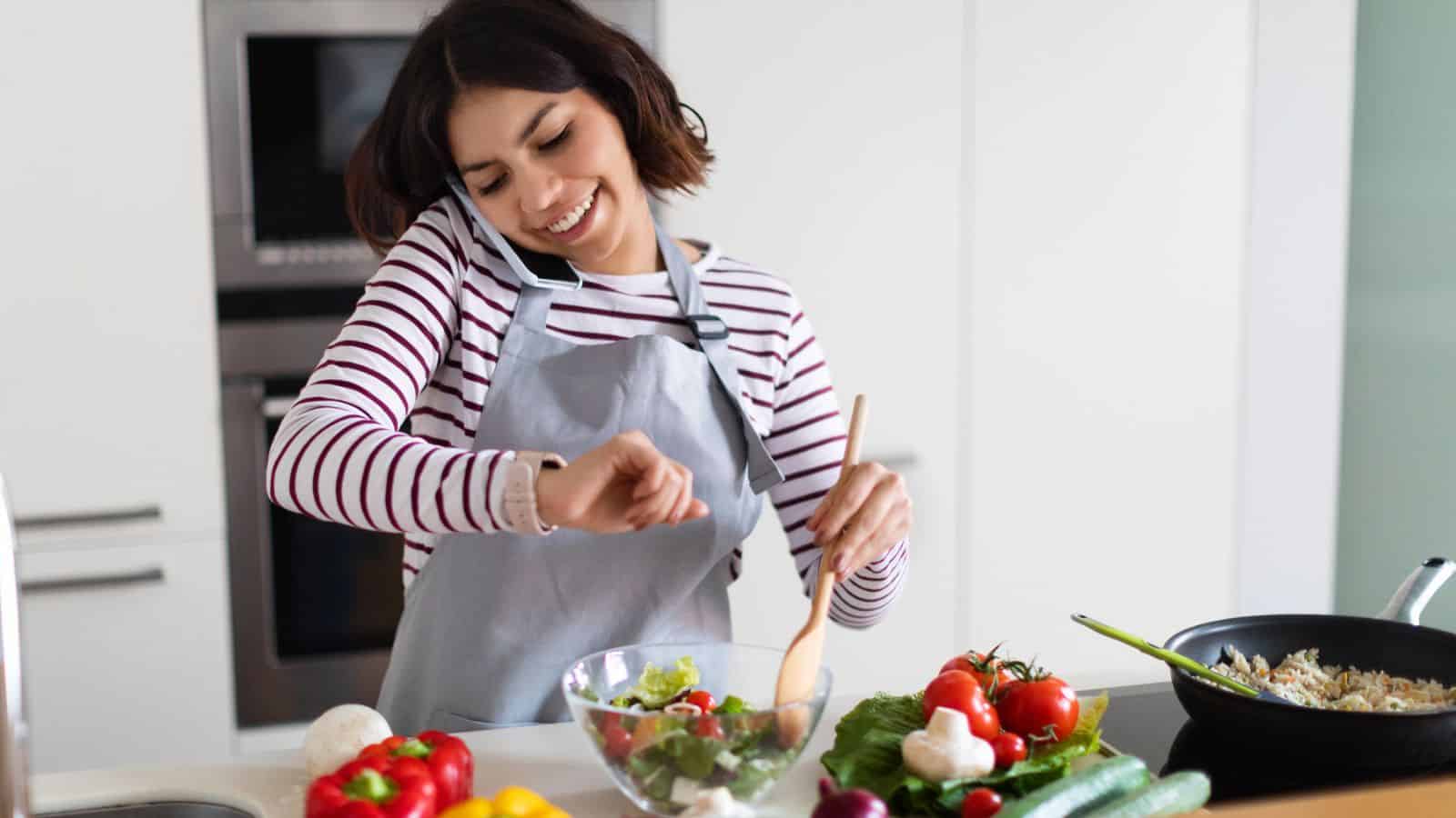 A woman wearing an apron talks on the phone while preparing a salad in a kitchen, checking her watch and surrounded by fresh vegetables.
