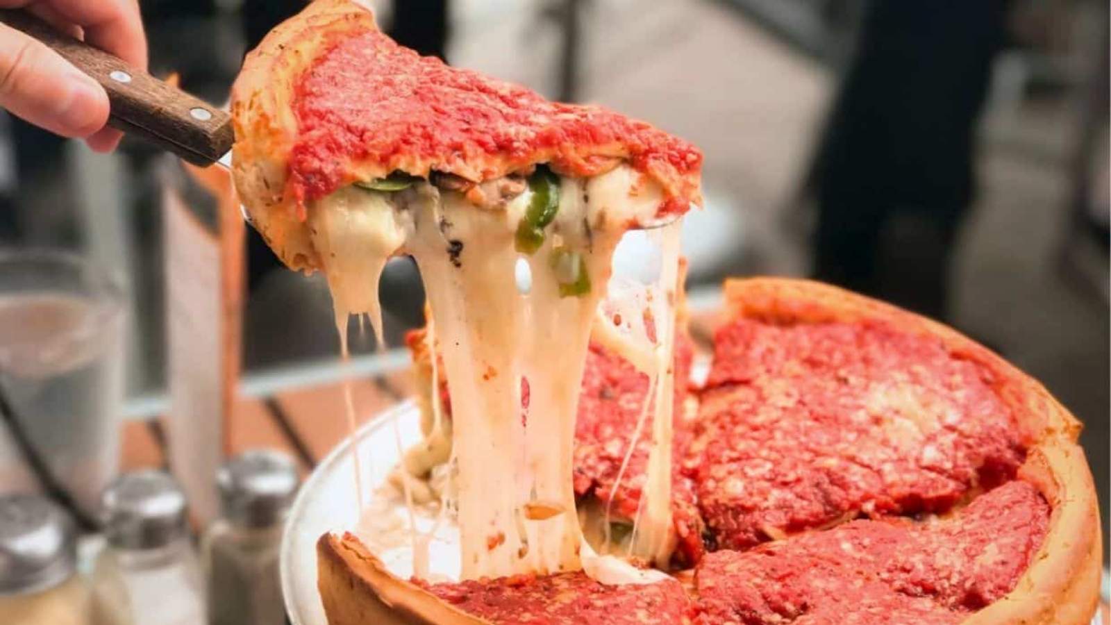 A slice of deep-dish pizza with melted cheese being lifted from a pan, showing a thick crust and tomato sauce topping.