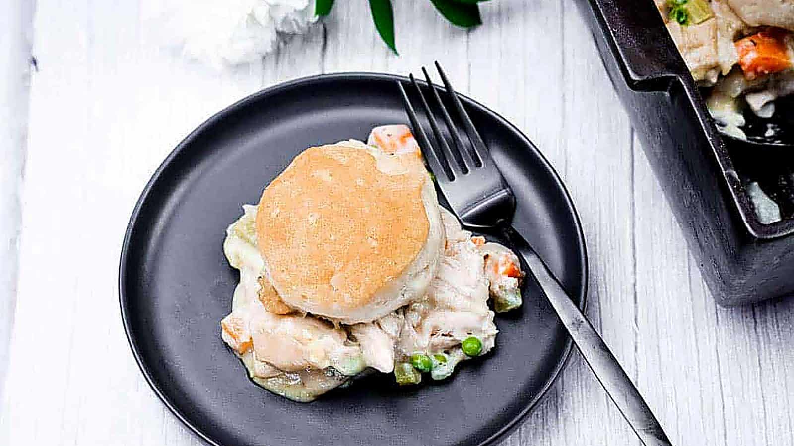 A black plate features a biscuit-topped serving of chicken pot pie, complete with chicken, peas, and carrots. A fork is placed on the plate, with a baking dish partially visible in the background.