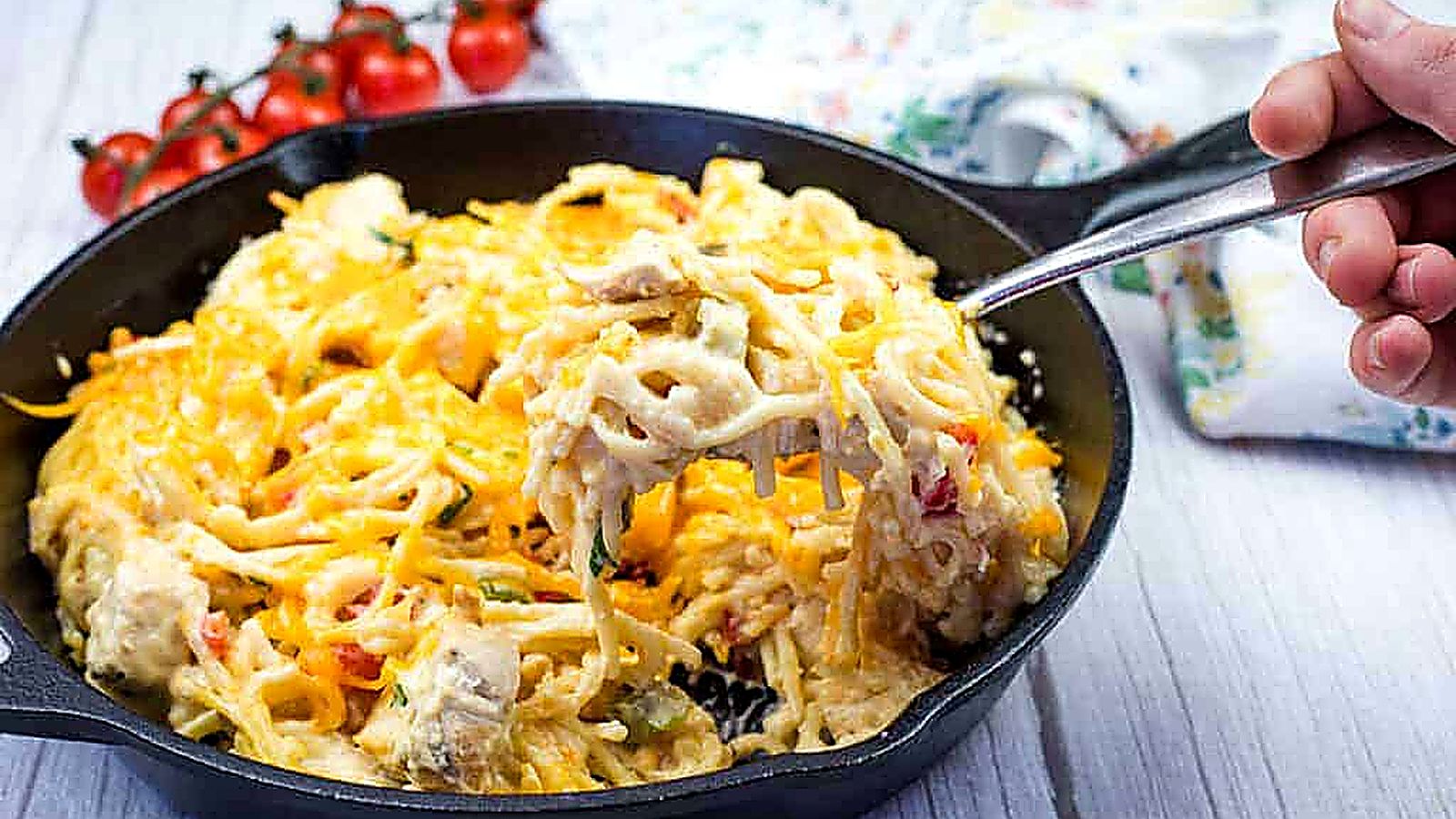 A hand holding a fork with a serving of chicken spaghetti from a cast-iron skillet.