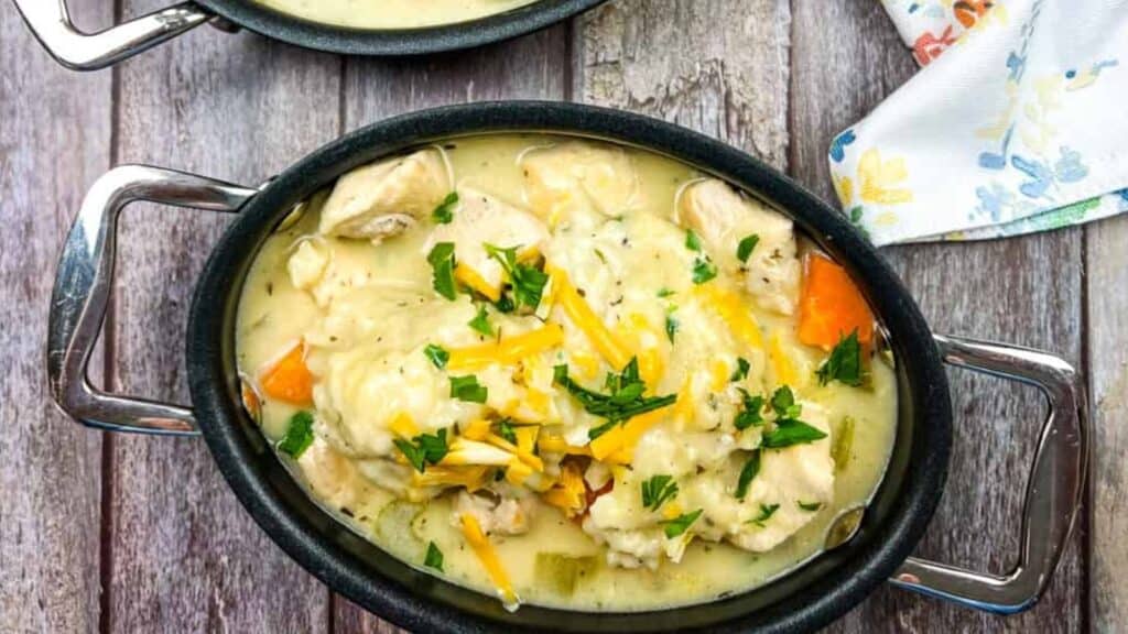 Oval bowl of creamy chicken and dumplings topped with shredded cheese and chopped parsley, with visible carrots and celery, on a wooden surface next to a floral napkin.