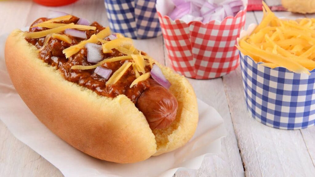 A chili cheese hot dog topped with shredded cheddar and diced onions, served on a paper liner with extra cups of chopped onions and shredded cheese&mdash;perfect for true hot dogs lovers.