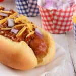 A chili cheese hot dog topped with shredded cheddar and diced onions, served on a paper liner with extra cups of chopped onions and shredded cheese—perfect for true hot dogs lovers.