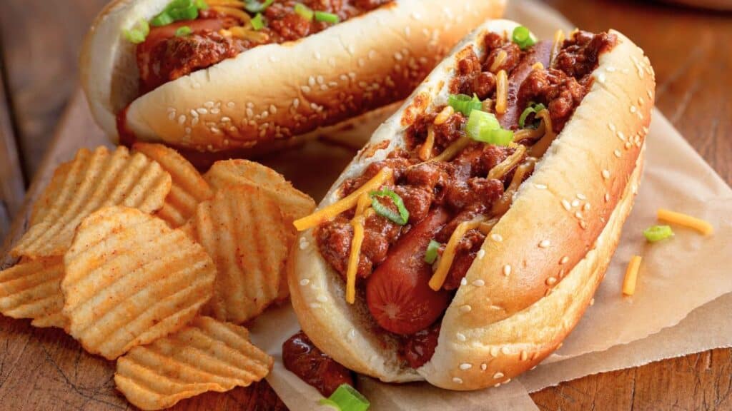 Two chili cheese hot dogs topped with shredded cheese and green onions, served on sesame seed buns, with a side of ridged potato chips on a wooden surface&mdash;because when it comes to game-day snacks, chili dog rules apply.