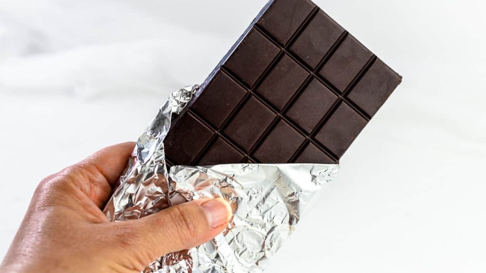 A hand holding a partially unwrapped dark chocolate bar in silver foil against a plain white background.