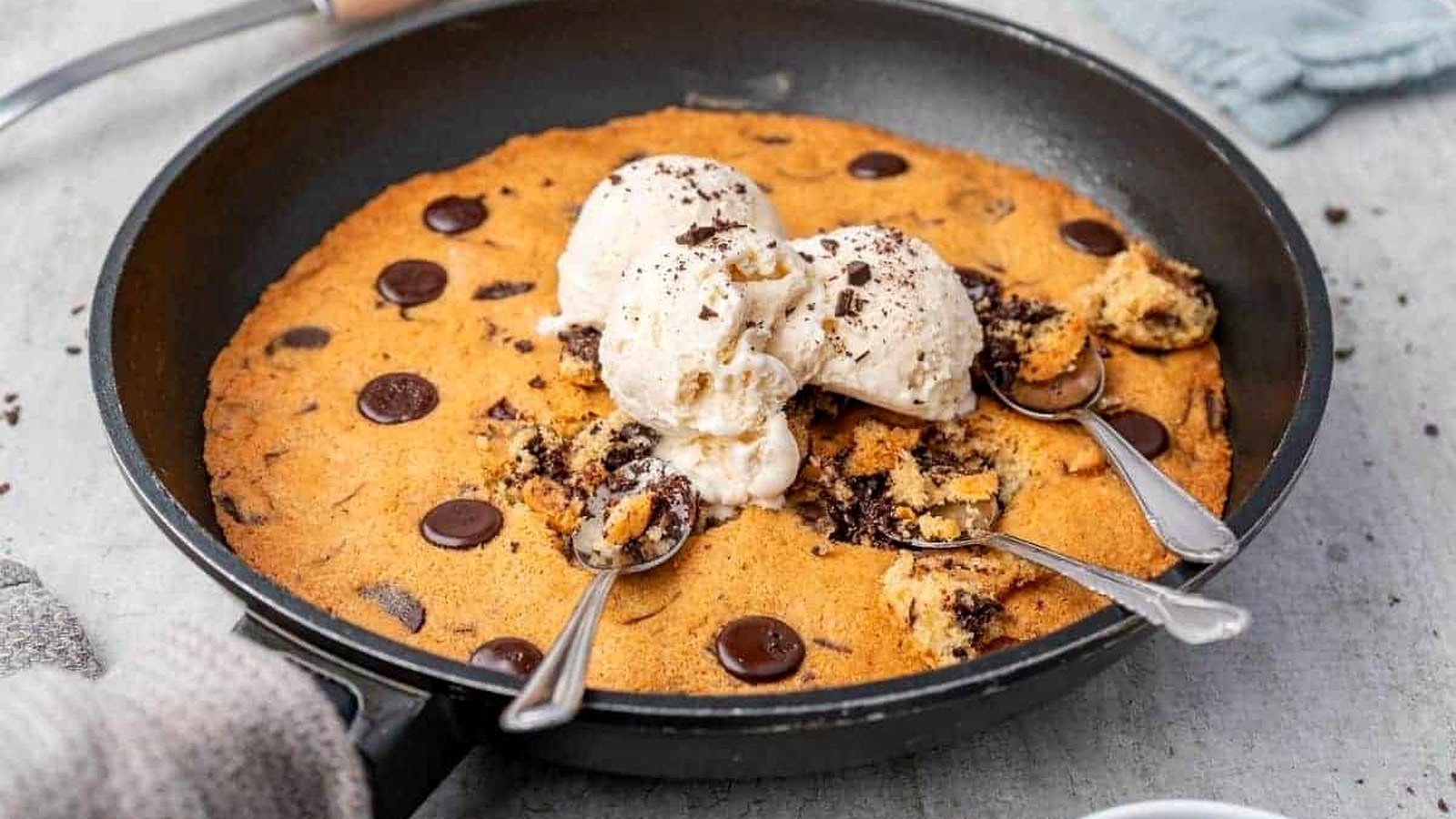 A slice of chocolate chip skillet cookie topped with a scoop of vanilla ice cream sits on a white plate with a spoon.