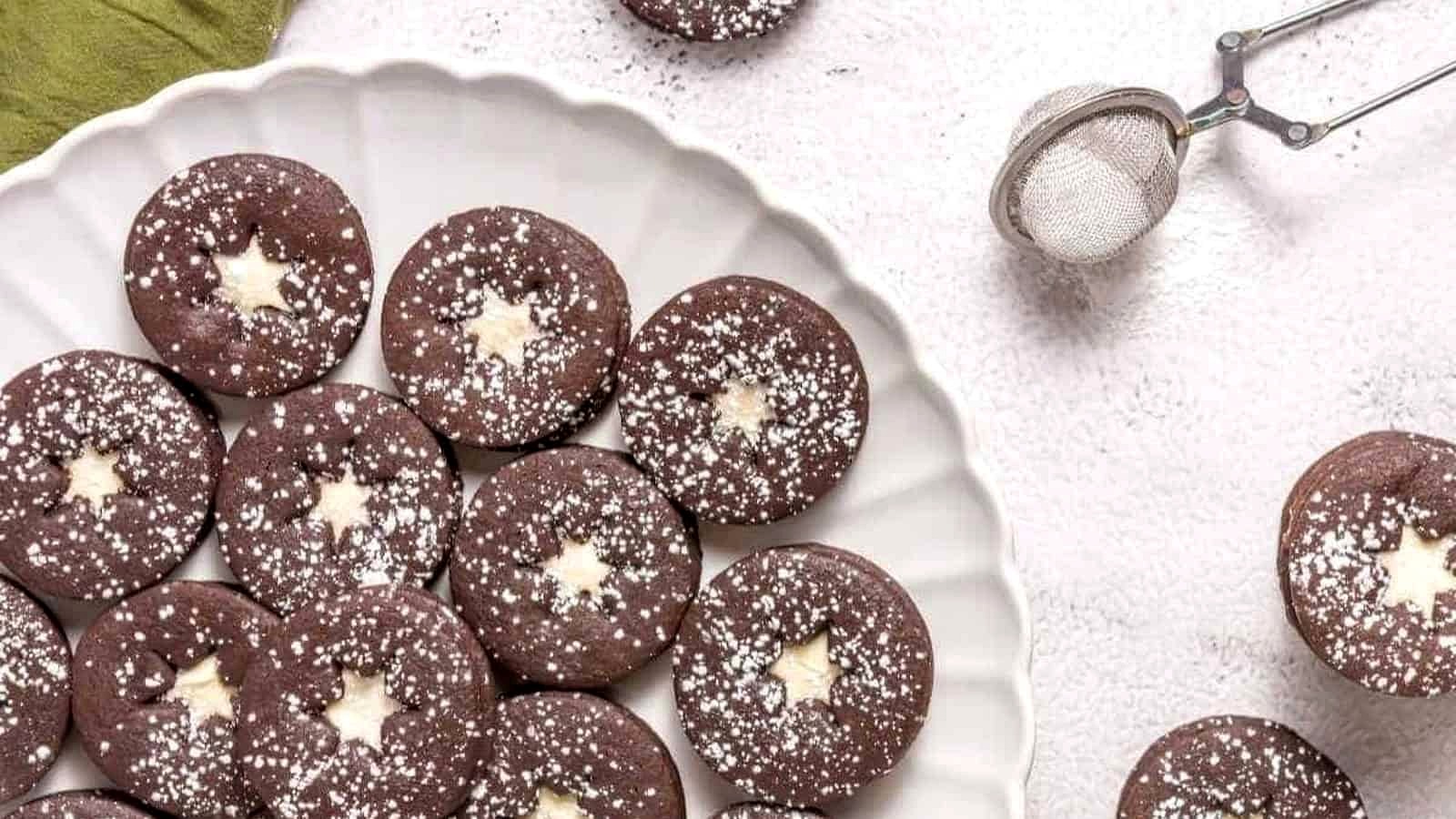 A plate of round chocolate cookies with star-shaped centers, dusted with powdered sugar. A metal sifter with powdered sugar is on the side.