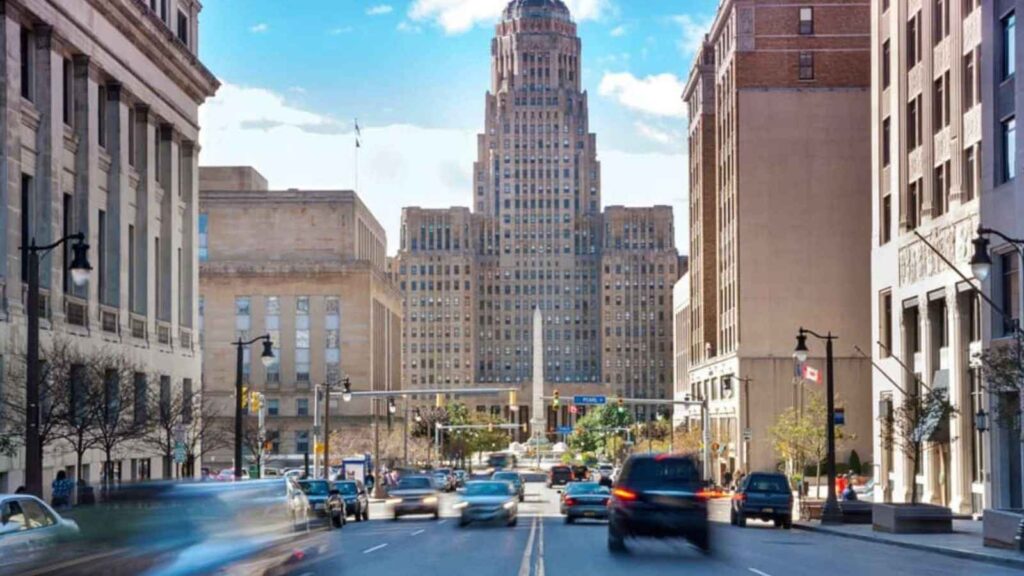A busy city street with cars and pedestrians, buzzing with Fourth of July travel, leads to a tall art deco building under a blue sky with scattered clouds.