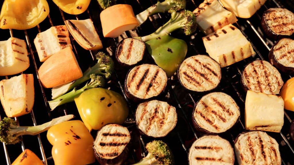 An assortment of sliced vegetables, including eggplant, bell pepper, broccoli, and squash, grilling on a barbecue and soaking up irresistible grilling flavors.